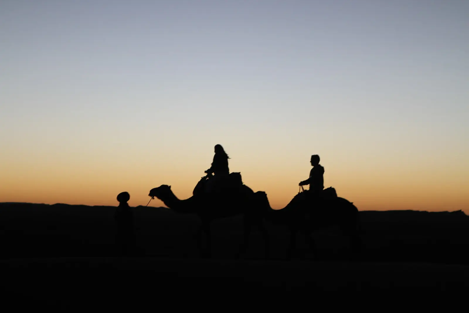 Two visitors riding camels in silhouette against a glowing orange sunset sky, led by a walking guide near Agadir