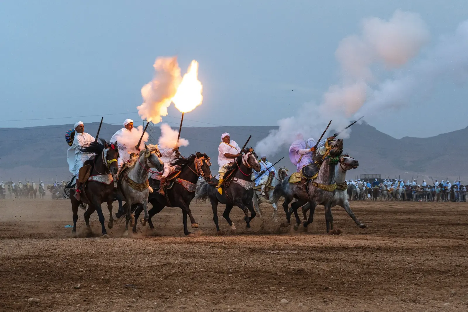 Horsemen in white robes charging across a dusty arena firing muskets with flame and gunpowder smoke during the Fantasia show