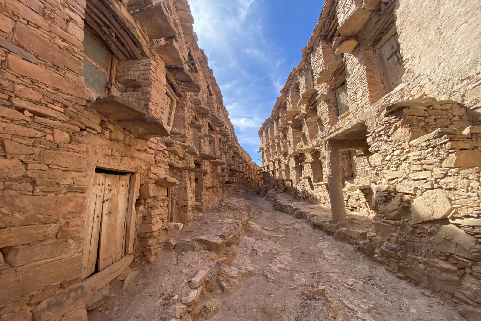 Interior lane of the Inoumar fortified granary lined with stone walls, wooden doors and grain-storage alcoves under a blue sky