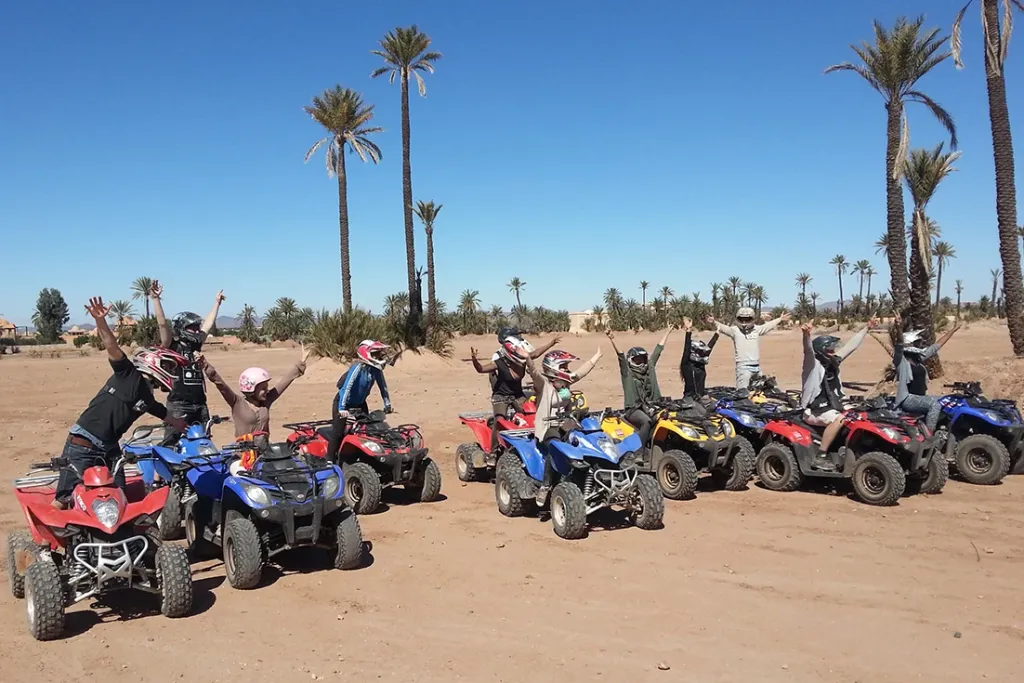A group of travelers on colorful quad bikes cheering with raised arms on a dusty trail lined with palm trees