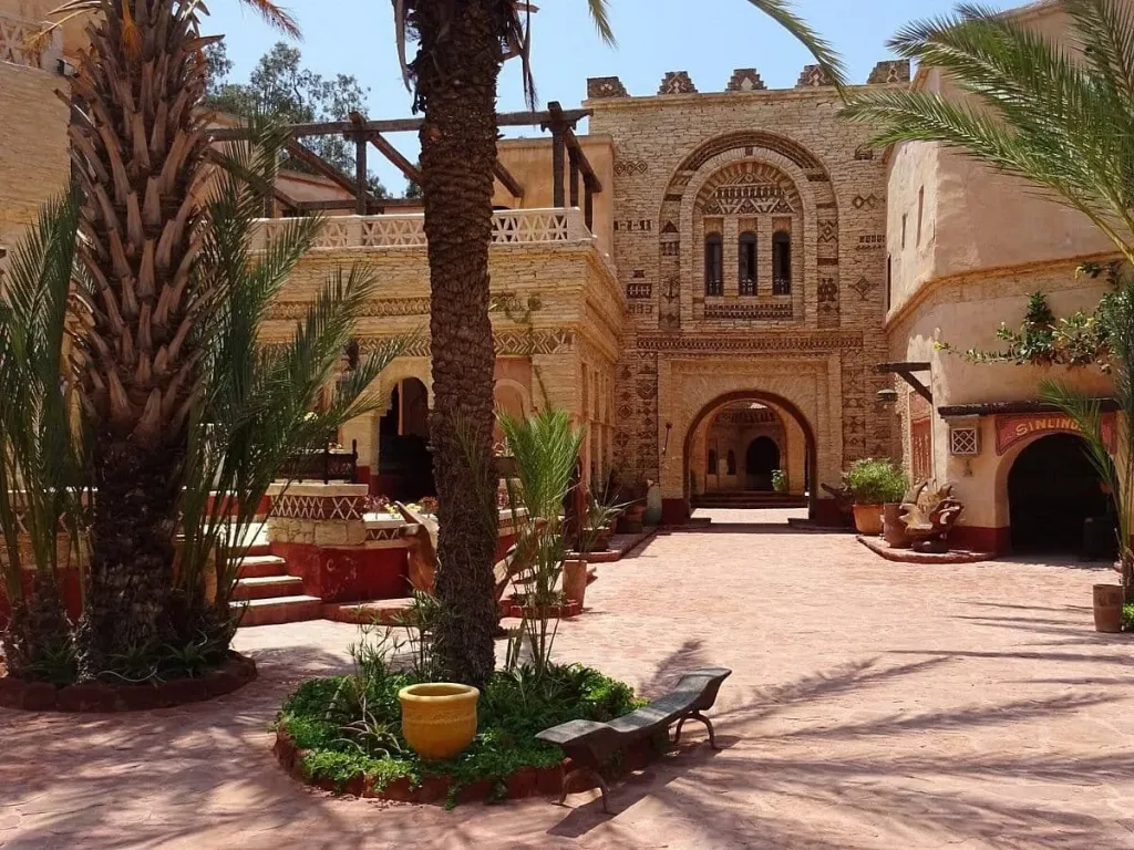 Sunlit courtyard of the Coco Polizzi medina in Agadir, with palm trees and a richly carved stone gateway