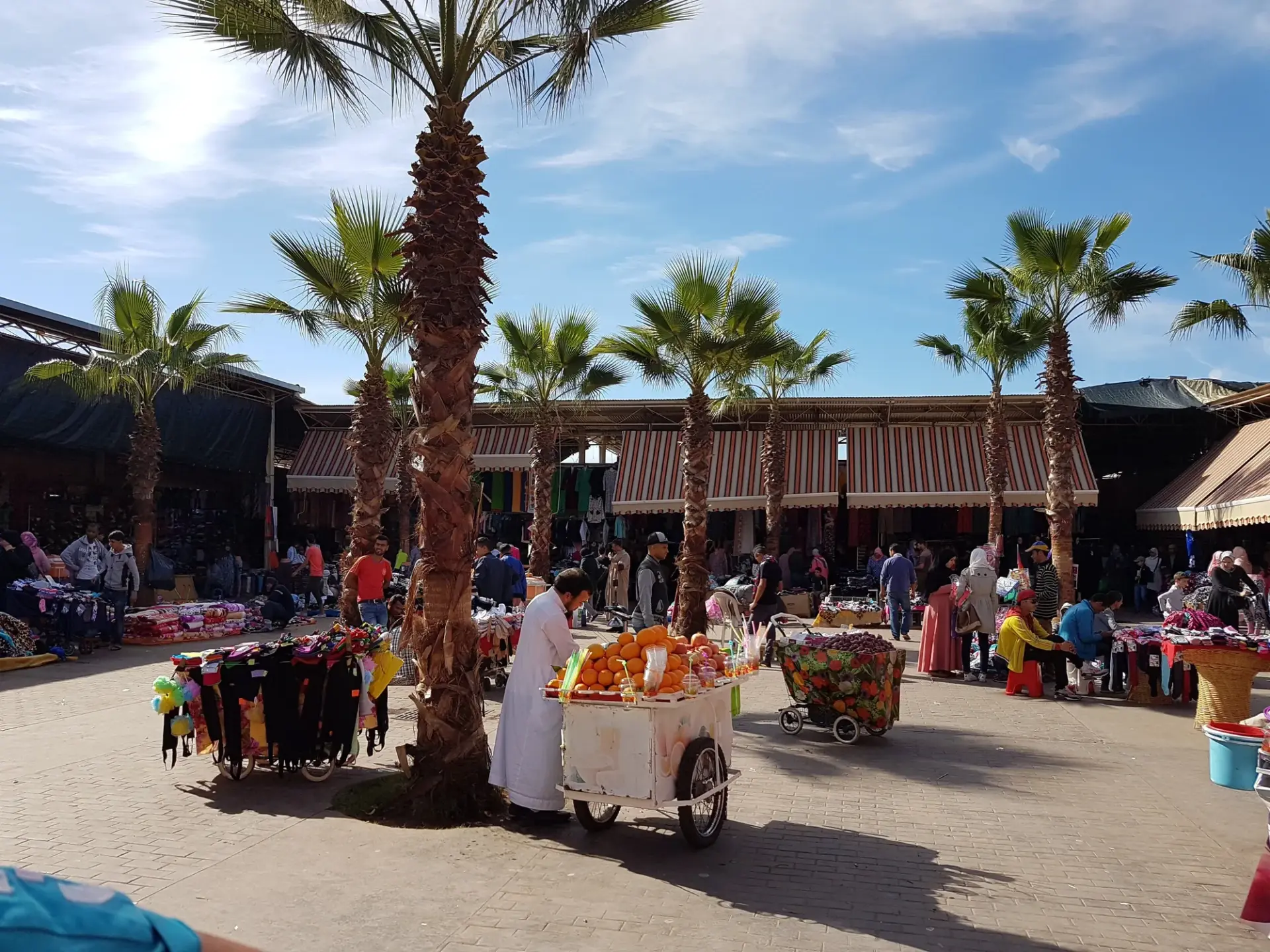 Busy square at Souk El Had in Agadir with palm trees, market stalls and an orange juice cart
