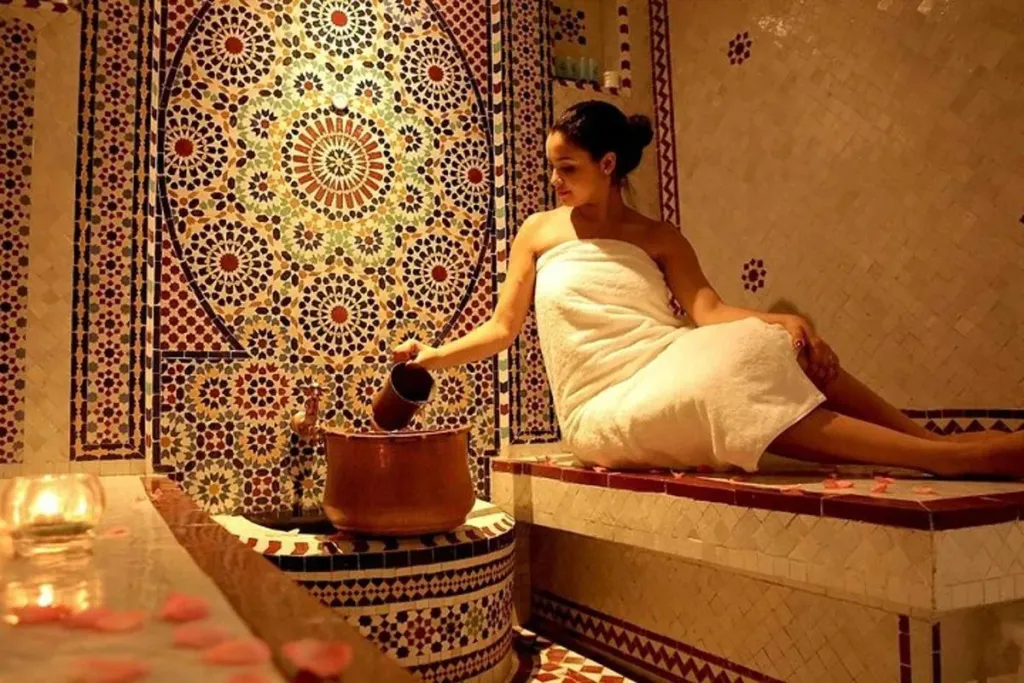 Woman wrapped in a towel pouring water at a mosaic-tiled fountain in a traditional Moroccan hammam