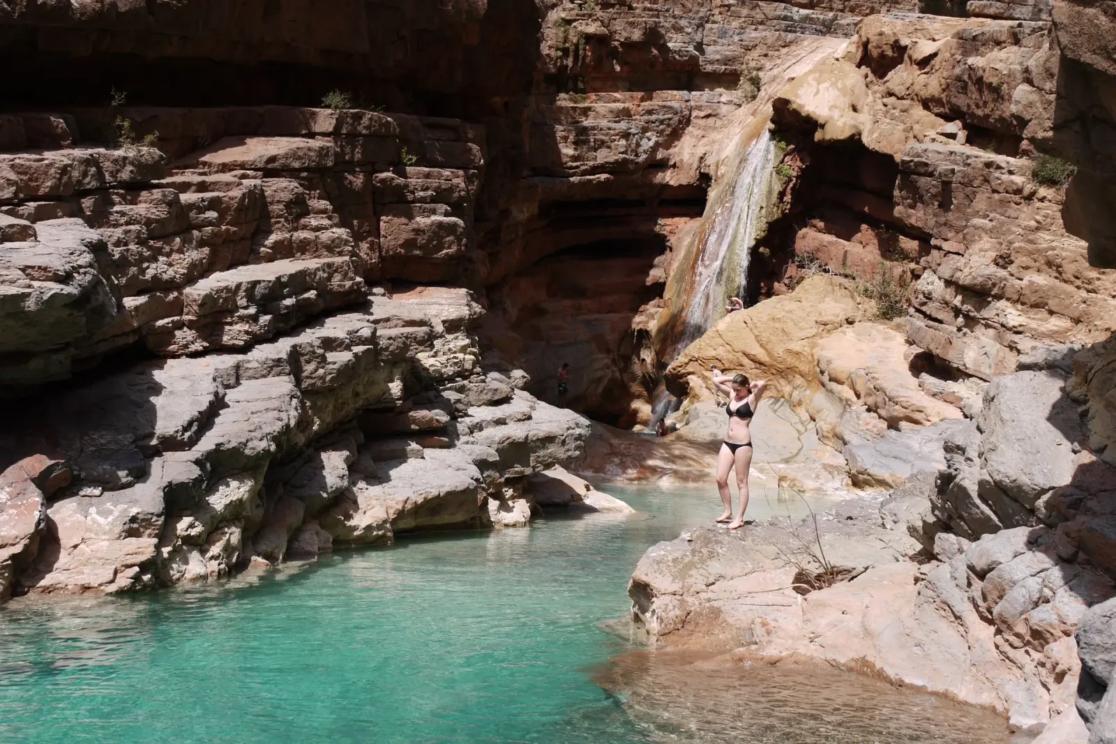 A traveler standing on the rocks beside a turquoise pool beneath a small waterfall in the palm gorge of Tizgui